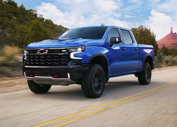 Blue Chevrolet pickup truck driving on a scenic road with hills in the background.