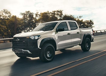 White Chevrolet pickup truck driving on a highway with blurred trees in the background.